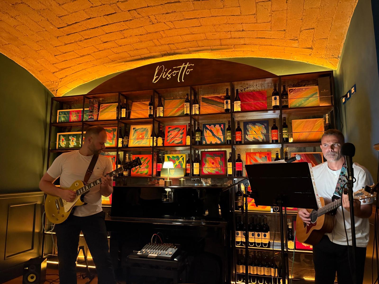 Il duo musicale elettroacustico AllYouCanSing si esibisce dal vivo al DiSotto di Roma, con chitarra acustica, chitarra elettrica e pianoforte, in un ambiente intimo con soffitto a volta e scaffali di bottiglie.
