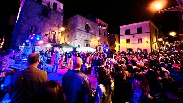 Una folla entusiasta assiste a uno spettacolo musicale live durante la Notte Rosa 2025 a Vitorchiano, con la band AllyoucanSing che si esibisce sotto luci colorate in un pittoresco borgo illuminato di notte